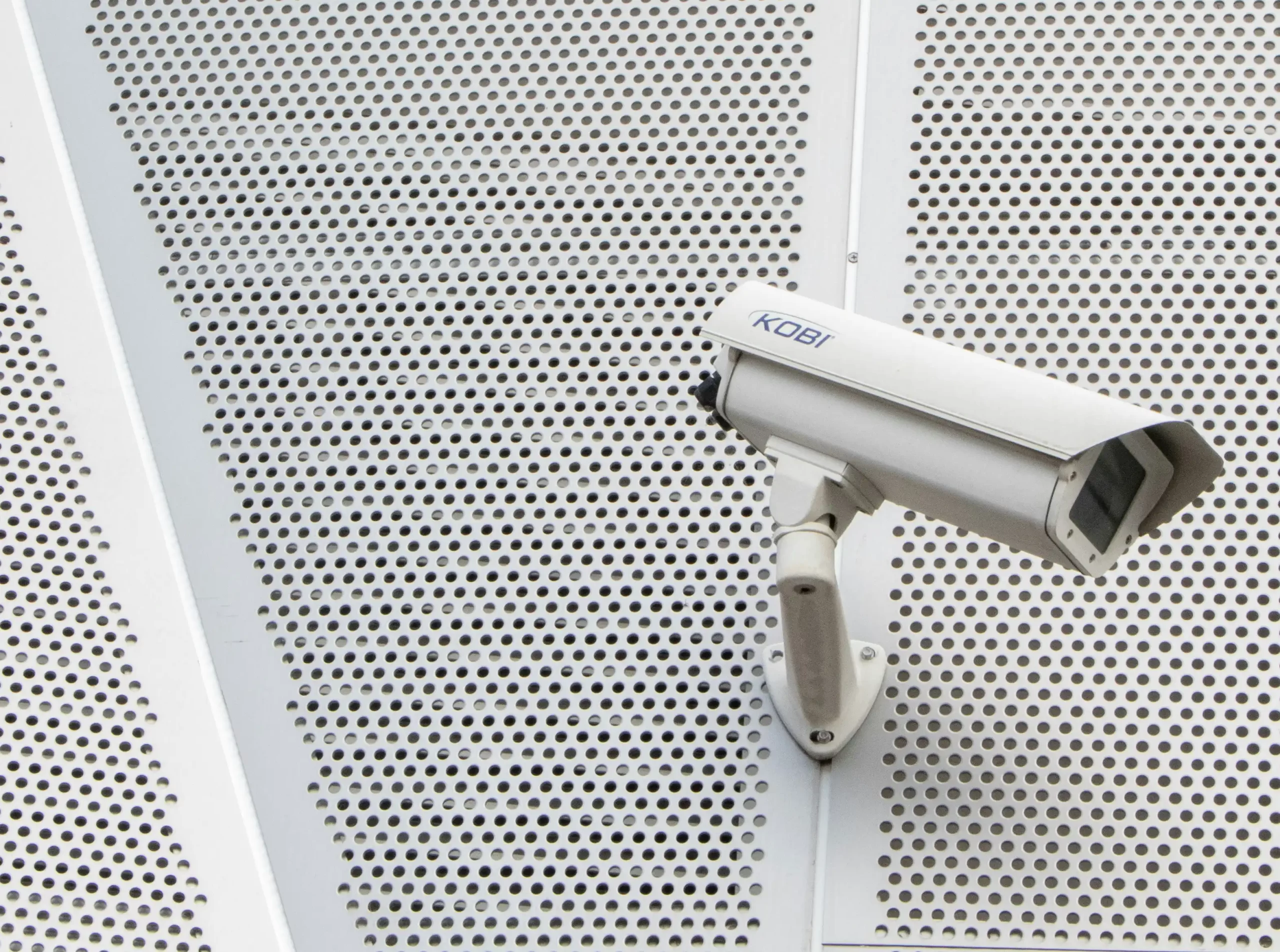 A white CCTV camera mounted on a perforated metal wall.